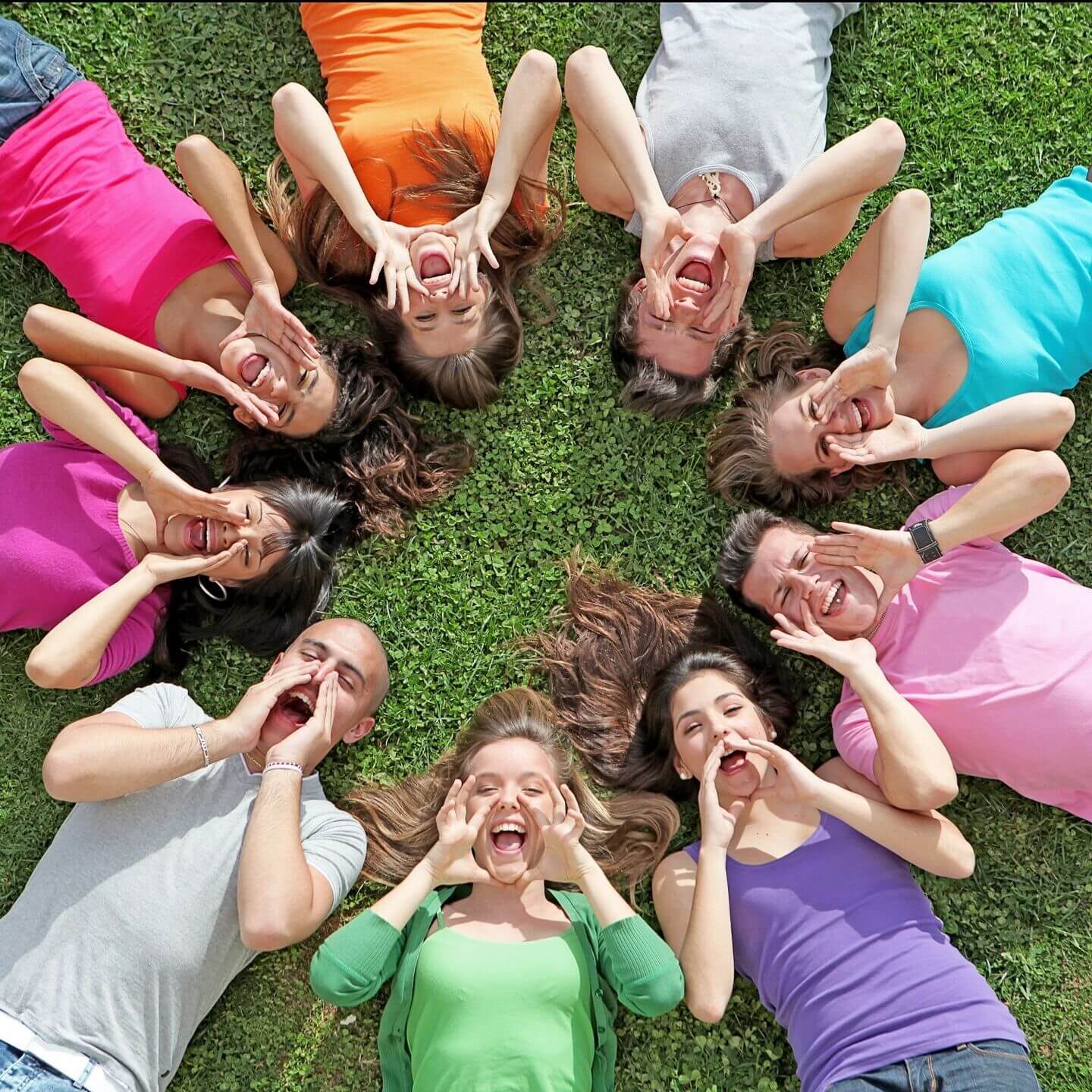 A circle of cheerful children lying on grass, making funny faces.